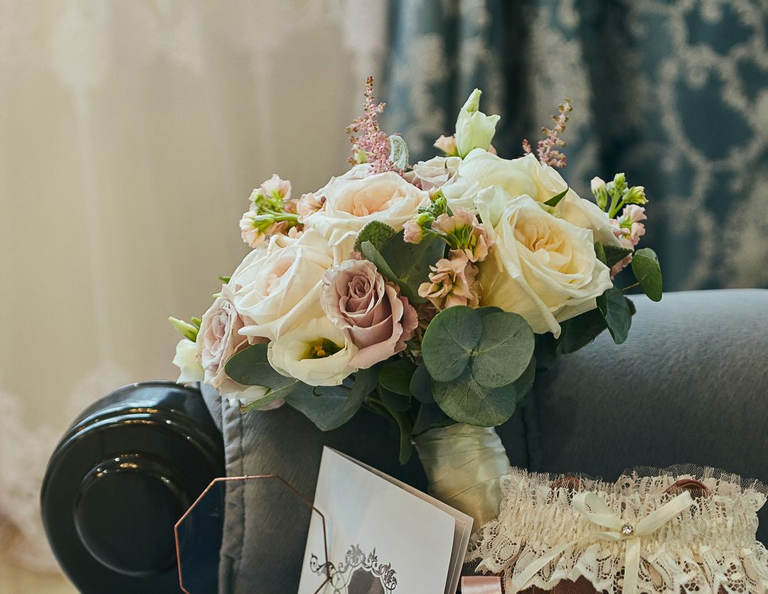 Ramo de rosas blancas y lila sobre un sillon gris junto a una tarjeta de invitacion.