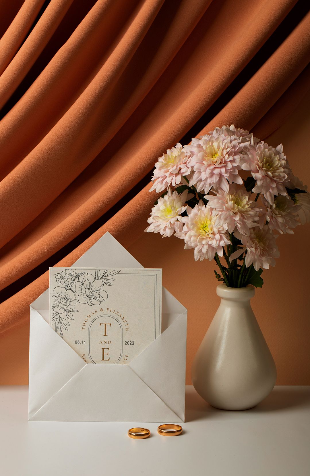 Wedding invitation card partially inside white envelope, with a beige vase holding light pink flowers and two gold wedding rings on a white surface.