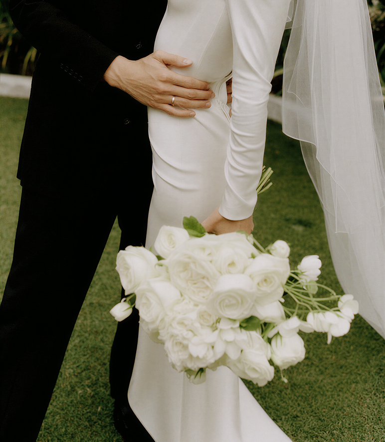 Close-up of a bride in a white dress and veil holding a bouquet of white flowers embraced by a groom in a black suit with a wedding ring on his hand.