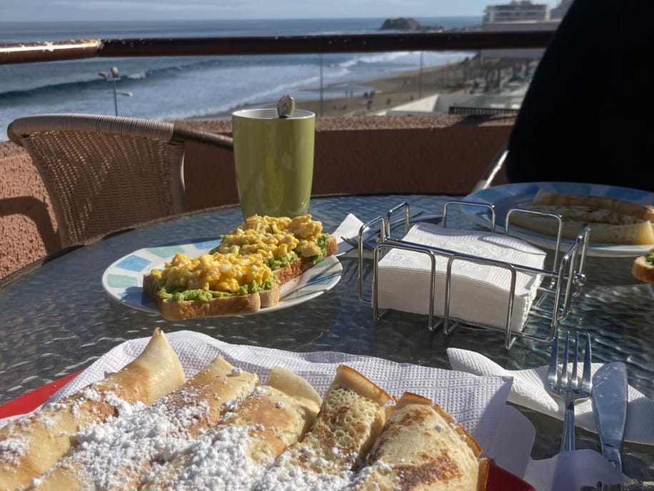 Desayuno de crepes y tostadas con palta sobre una mesa de terraza, con la playa y el mar de fondo